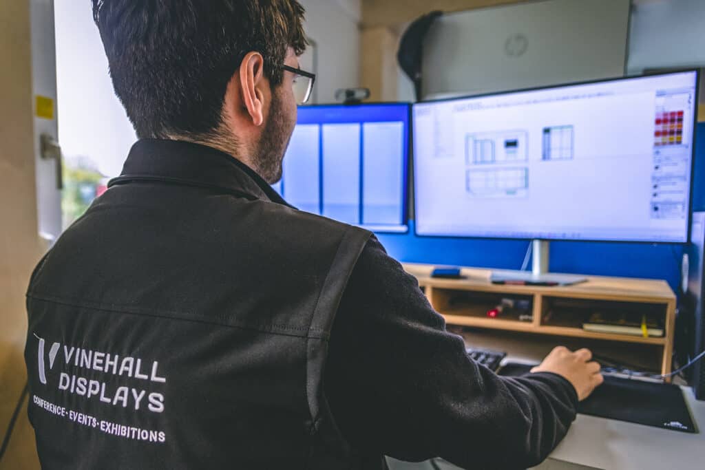 a man wearing a Vinehall Displays jacket works at a computer to plan a project budget